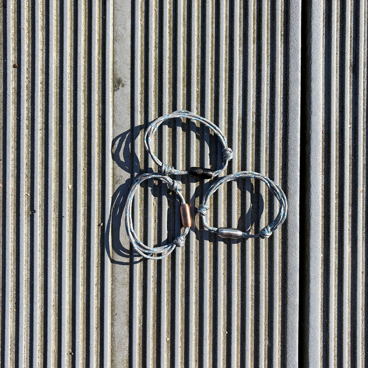 Drei größenverstellbare Baltic Sea Bracenets, gefertigt aus alten Fischernetzen in Grau mit Punkten in Blau und Weiß und mit Bajonettverschluss in Roségold, Schwarz oder Silber liegen am Boden. 