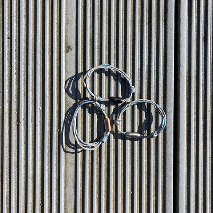 Drei größenverstellbare Baltic Sea Bracenets, gefertigt aus alten Fischernetzen in Grau mit Punkten in Blau und Weiß und mit Bajonettverschluss in Roségold, Schwarz oder Silber liegen am Boden. 