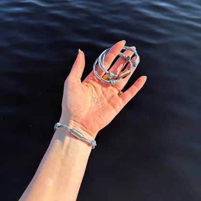 Person hält drei größenverstellbare Baltic Sea Bracenet, gefertigt aus alten Fischernetzen in Grau mit Punkten in Blau und Weiß und mit Bajonettverschluss in Roségold, Schwarz und Silber in der Hand über dem Wasser. 