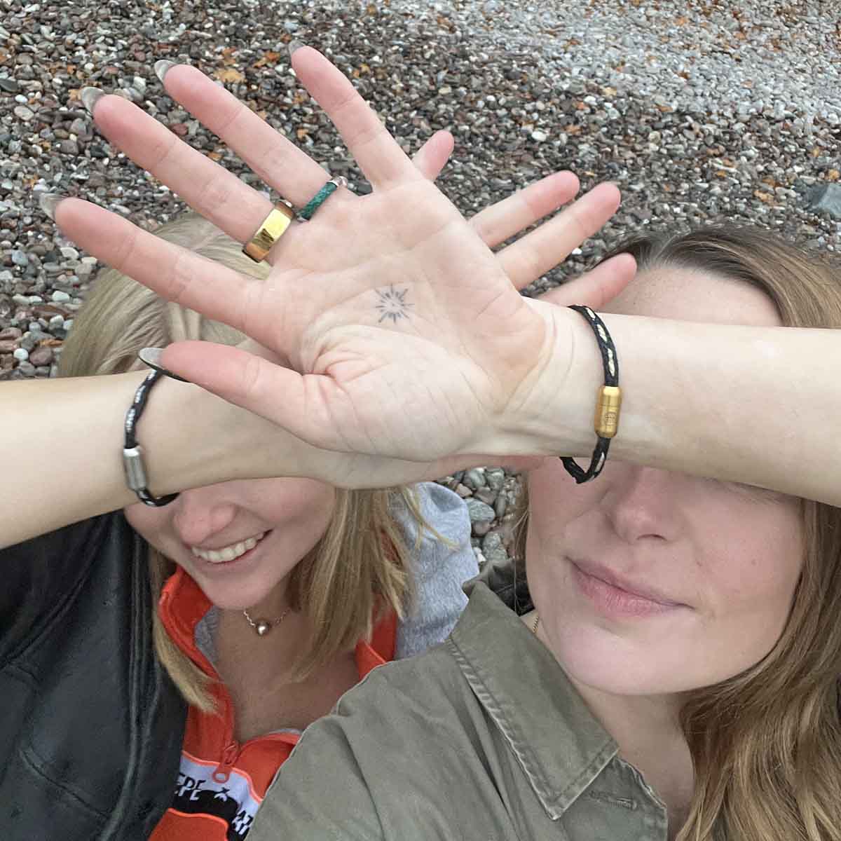 Zwei Personen strecken die Hände in die Kamera und vor ihr Gesicht. An den Handgelenk trägt eine Person das BRACENET Glam Edition Armband aus schwarzem Fischernetz mit goldenem Faden durchzogen und goldenem Magnetverschluss und die andere das BRACENET Glam Edition Armband aus schwarzem Fischernetz mit silbernem Faden durchzogen und silbernem Magnetverschluss.