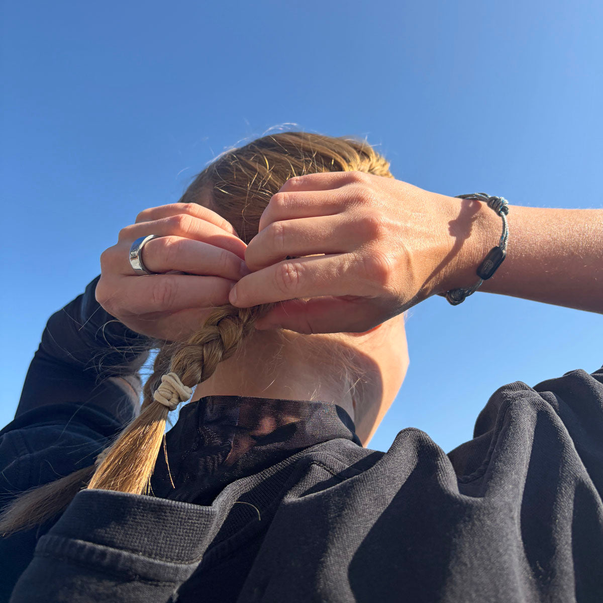 Person trägt das größenverstellbare Baltic Sea Bracenet, gefertigt aus alten Fischernetzen in Grau mit Punkten in Blau und Weiß und mit Bajonettverschluss in Schwarz am Handgelenk und fasst sich in die Haare. 