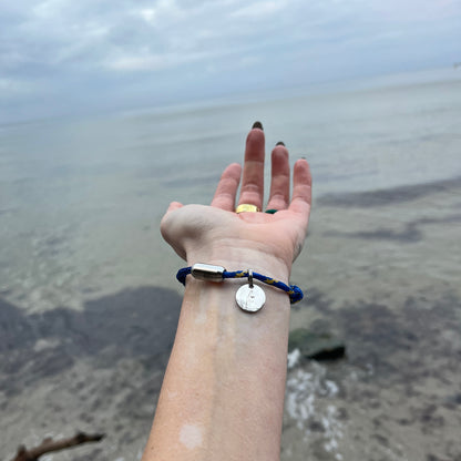 Person trägt das BRACENET Upcycling Armband Insel Sylt II aus alten Fischernetzen in dunkelblau und blau-gelb, mit einem silbernen Magnetverschluss und Anhänger mit einer Inselgravur von Sylt am Handgelenk und hält die Hand vor das Meer. 