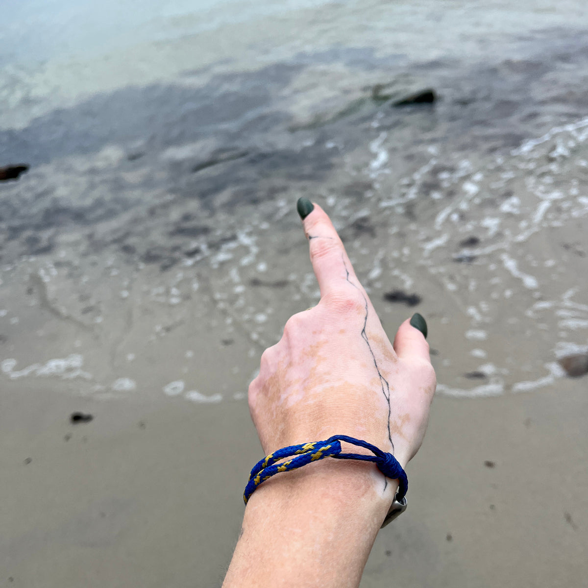 Person trägt das BRACENET Upcycling Armband Insel Sylt II aus alten Fischernetzen in dunkelblau und blau-gelb, mit einem silbernen Magnetverschluss und Anhänger mit einer Inselgravur von Sylt am Handgelenk und zeigt auf das Meer. 