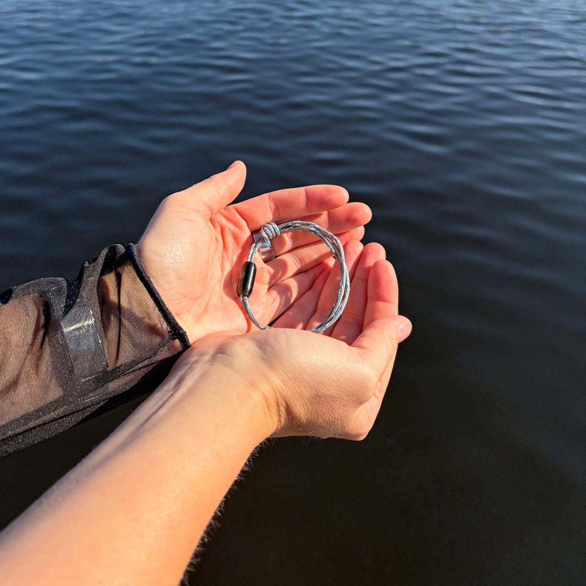Person hälz das größenverstellbare Baltic Sea Bracenet, gefertigt aus alten Fischernetzen in Grau mit Punkten in Blau und Weiß und mit Bajonettverschluss in Schwarz in den Händen über dem Wasser. 