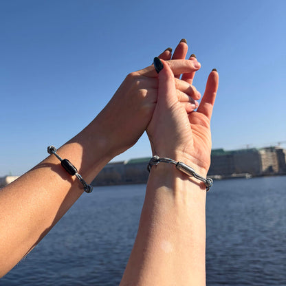 Zwei Personen halten sich an den Händen und tragen das größenverstellbare Baltic Sea Bracenet, gefertigt aus alten Fischernetzen in Grau mit Punkten in Blau und Weiß und mit Bajonettverschluss in Schwarz und Silber. 