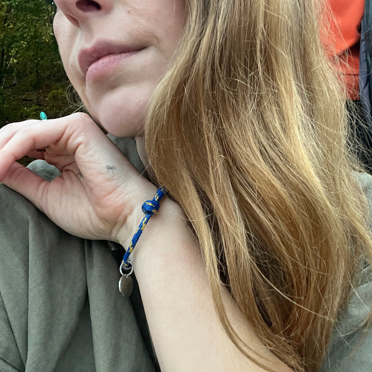 Person trägt das BRACENET Upcycling Armband Insel Sylt II aus alten Fischernetzen in dunkelblau und blau-gelb, mit einem silbernen Magnetverschluss und Anhänger mit einer Inselgravur von Sylt am Handgelenk und hebt die Hand ans Kinn.