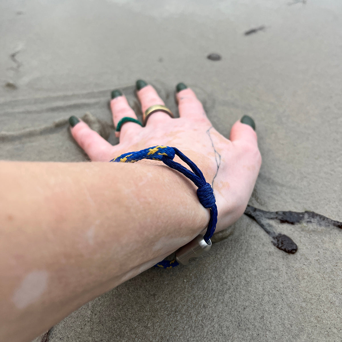 Person trägt das BRACENET Upcycling Armband Insel Sylt II aus alten Fischernetzen in dunkelblau und blau-gelb, mit einem silbernen Magnetverschluss und Anhänger mit einer Inselgravur von Sylt am Handgelenk und fährt mit der Hand durch den Sand. 