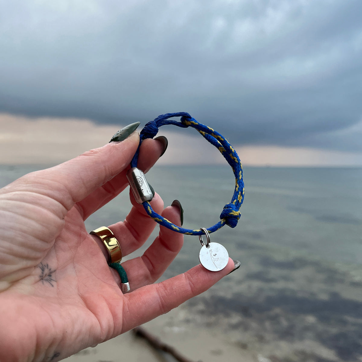 Person hält das BRACENET Upcycling Armband Insel Sylt II aus alten Fischernetzen in dunkelblau und blau-gelb, mit einem silbernen Magnetverschluss und Anhänger mit einer Inselgravur von Sylt in der Hand. Im Hintergrund sieht man das Meer. 
