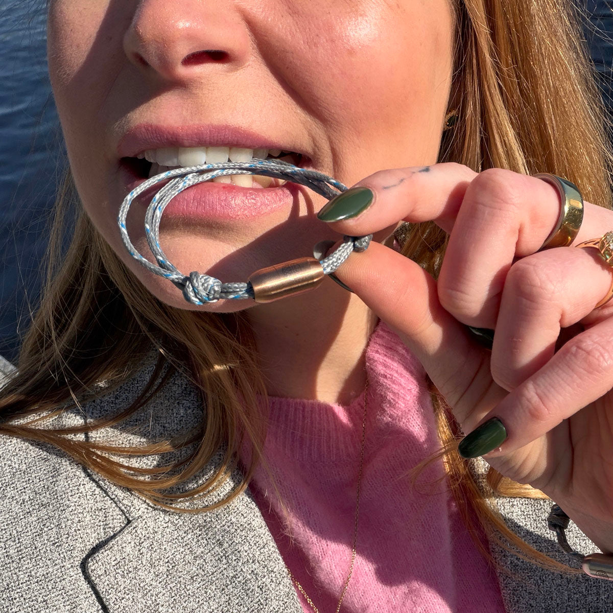 Person beißt auf das größenverstellbare Baltic Sea Bracenet, gefertigt aus alten Fischernetzen in Grau mit Punkten in Blau und Weiß und mit Bajonettverschluss in Roségold.