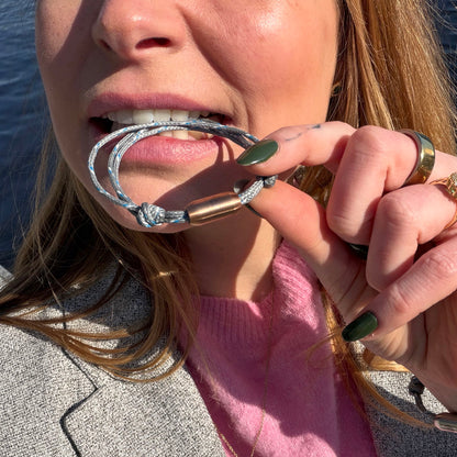 Person beißt auf das größenverstellbare Baltic Sea Bracenet, gefertigt aus alten Fischernetzen in Grau mit Punkten in Blau und Weiß und mit Bajonettverschluss in Roségold.