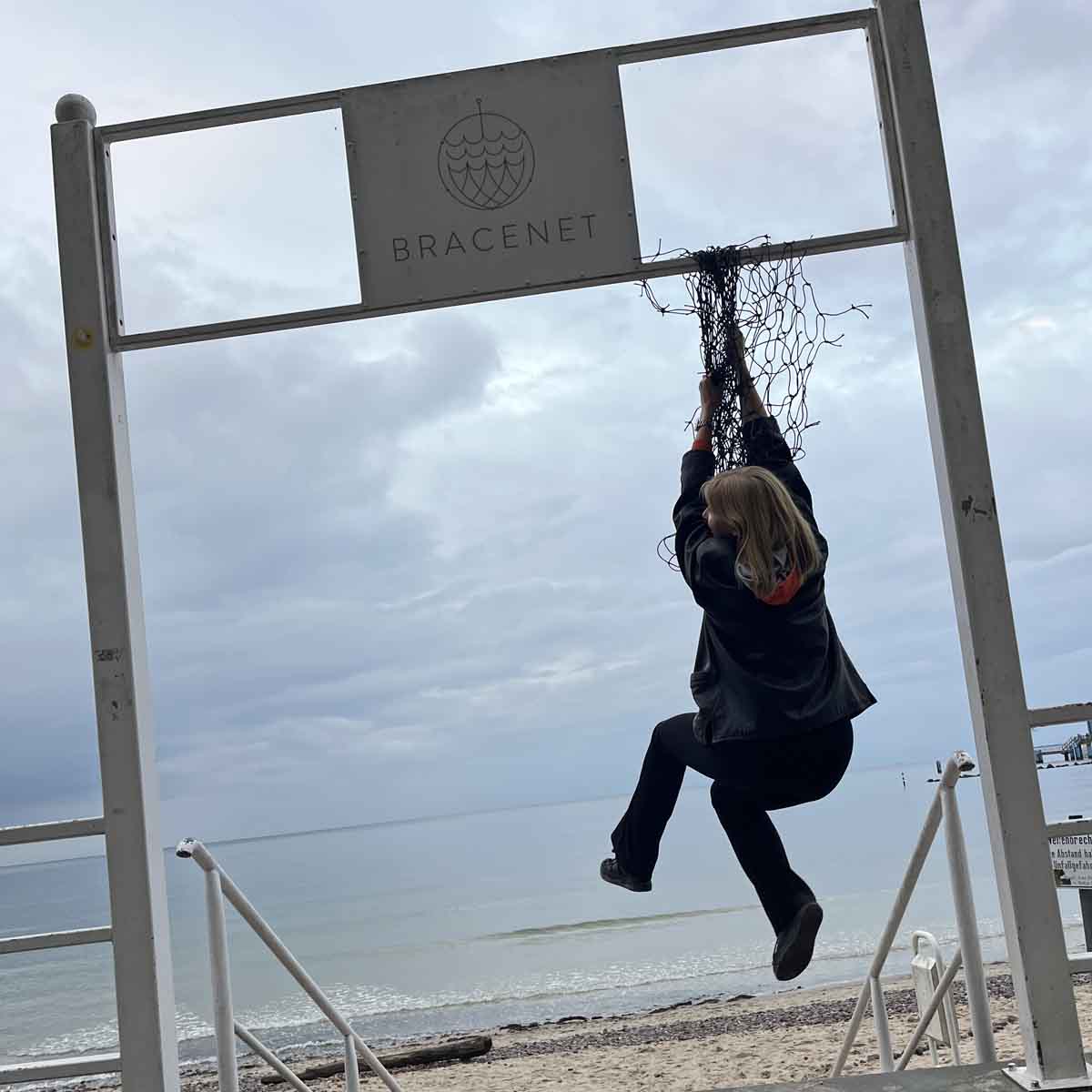 Eine Person hängt an einem Fischernetz von einem Schild. Auf dem Schild ist das BRACENET Logo zusehen. 