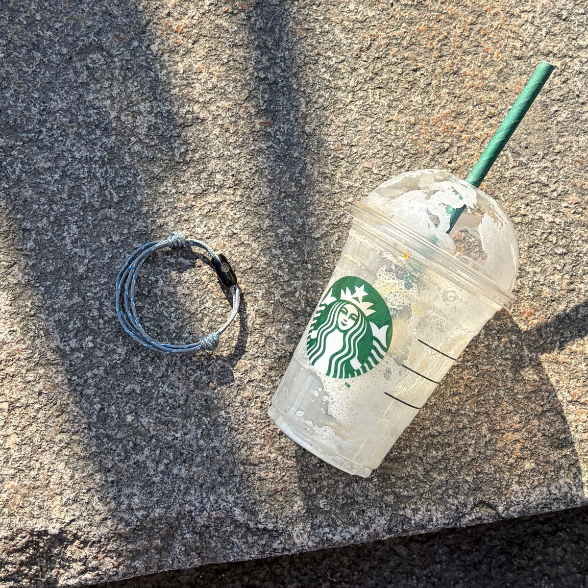 Das größenverstellbare Baltic Sea Bracenet, gefertigt aus alten Fischernetzen in Grau mit Punkten in Blau und Weiß und mit Bajonettverschluss in Schwarz liegt auf dem Boden neben einem Plastikbecher. 