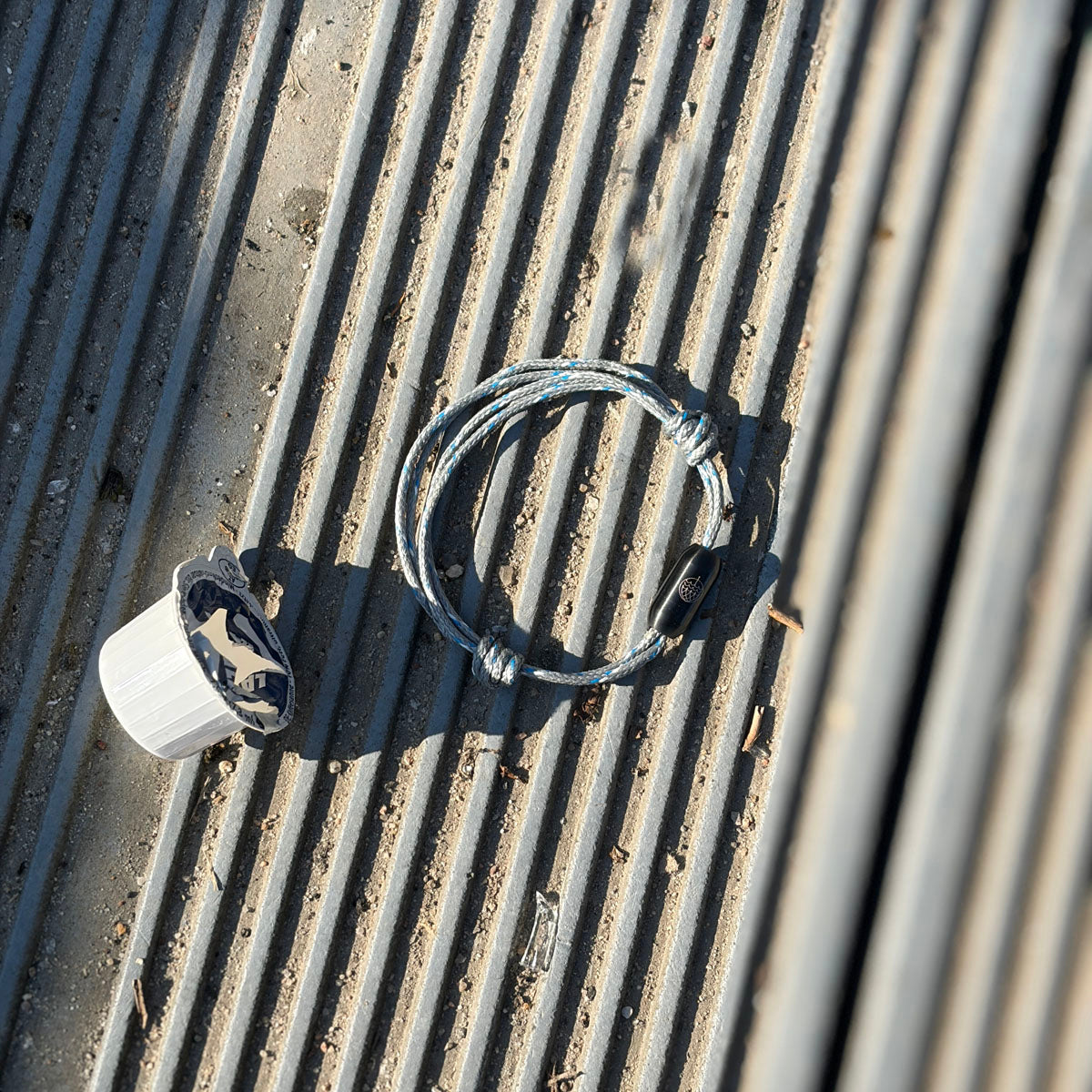 Das größenverstellbare Baltic Sea Bracenet, gefertigt aus alten Fischernetzen in Grau mit Punkten in Blau und Weiß und mit Bajonettverschluss in Schwarz liegt am Boden neben Plastikmüll.