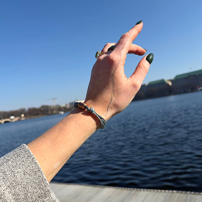 Person hat das as größenverstellbare Baltic Sea Bracenet, gefertigt aus alten Fischernetzen in Grau mit Punkten in Blau und Weiß und mit Bajonettverschluss in Roségold am Handgelenk und zeigt auf das Wasser. 