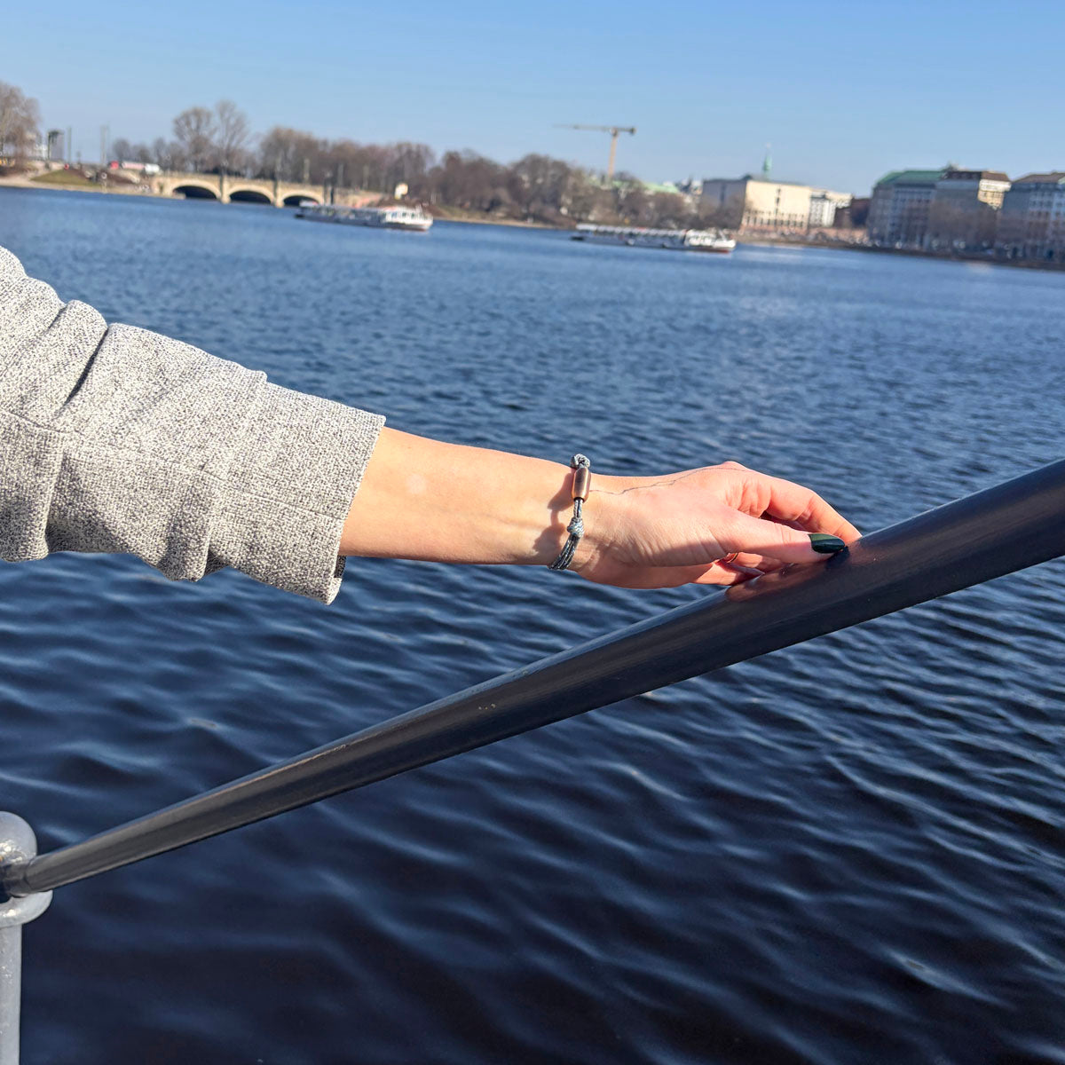 Person trägt das größenverstellbare Baltic Sea Bracenet, gefertigt aus alten Fischernetzen in Grau mit Punkten in Blau und Weiß und mit Bajonettverschluss in Roségold am Handgelenk und hält sich am Geländer fest. 