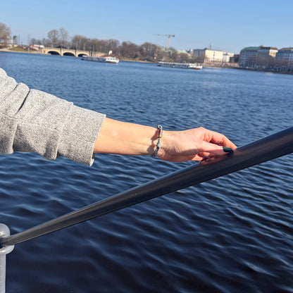 Person trägt das größenverstellbare Baltic Sea Bracenet, gefertigt aus alten Fischernetzen in Grau mit Punkten in Blau und Weiß und mit Bajonettverschluss in Roségold am Handgelenk und hält sich am Geländer fest. 