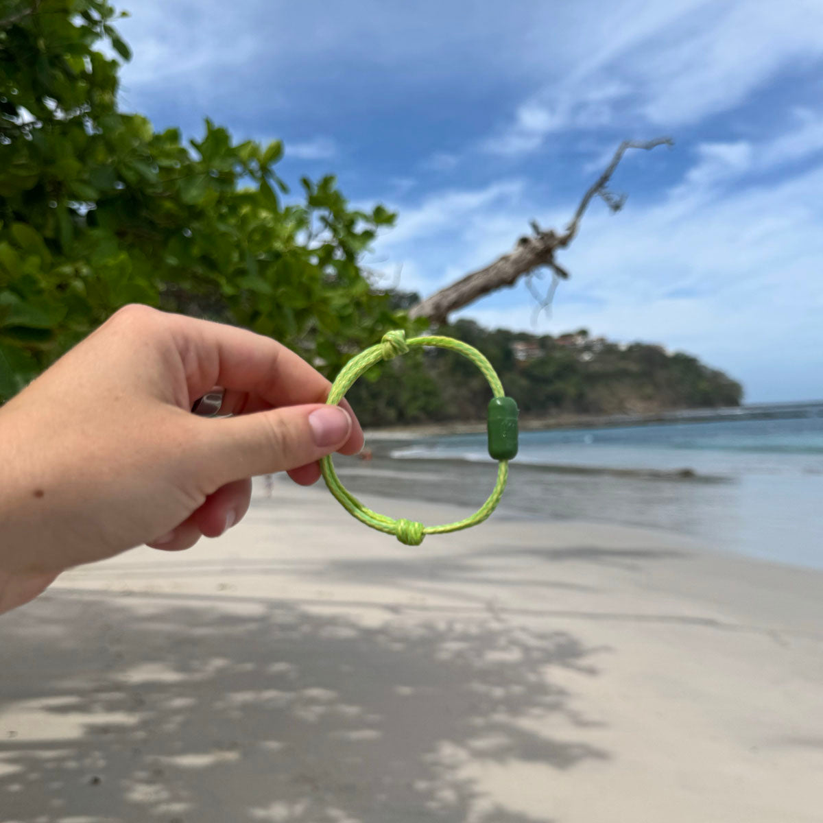 Person hält das Juni Bracenet Bismarck Sea aus Fischernetz in Hellgrün mit Magnetverschluss aus eingeschmolzenen Fischernetzen in Grün zwischen den Fingern in der Hand am Strand. 