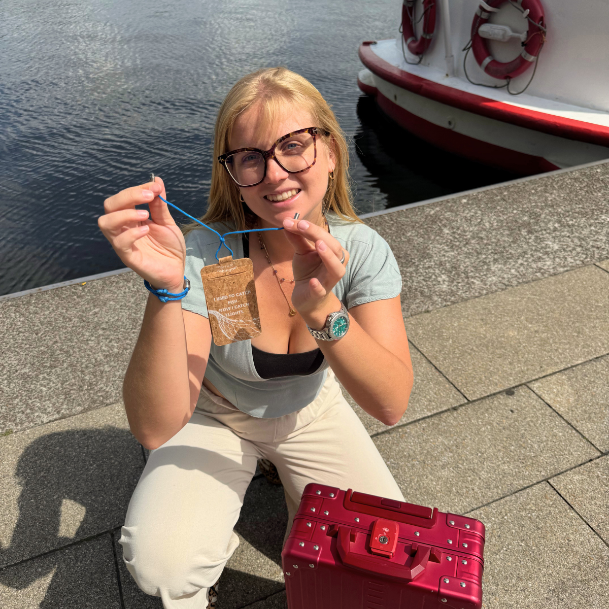 Person hat den BRACENET Kofferanhänger Arctic Ocean aus Kork mit Aufdruck der Aufschrift "I USED TO CATCH FISH. NOW I CATCH FLIGHTS." mit einer Schlaufe aus altem Fischernetz in Blau mit Drehverschluss in Silber geöffnet in der Hand. 