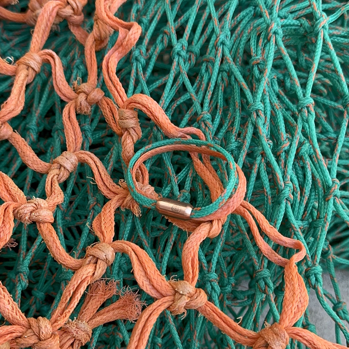 August Bracenet Phuket Armband aus Fischernetz mit einem Strang Fischernetz in Grün mit Highlights in Orange, einem Strang Fischernetz in Orange und einem Magnetverschluss in Roségold liegt auf Fischernetzen.