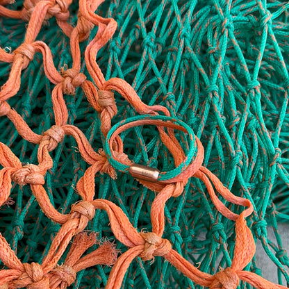 August Bracenet Phuket Armband aus Fischernetz mit einem Strang Fischernetz in Grün mit Highlights in Orange, einem Strang Fischernetz in Orange und einem Magnetverschluss in Roségold liegt auf Fischernetzen.