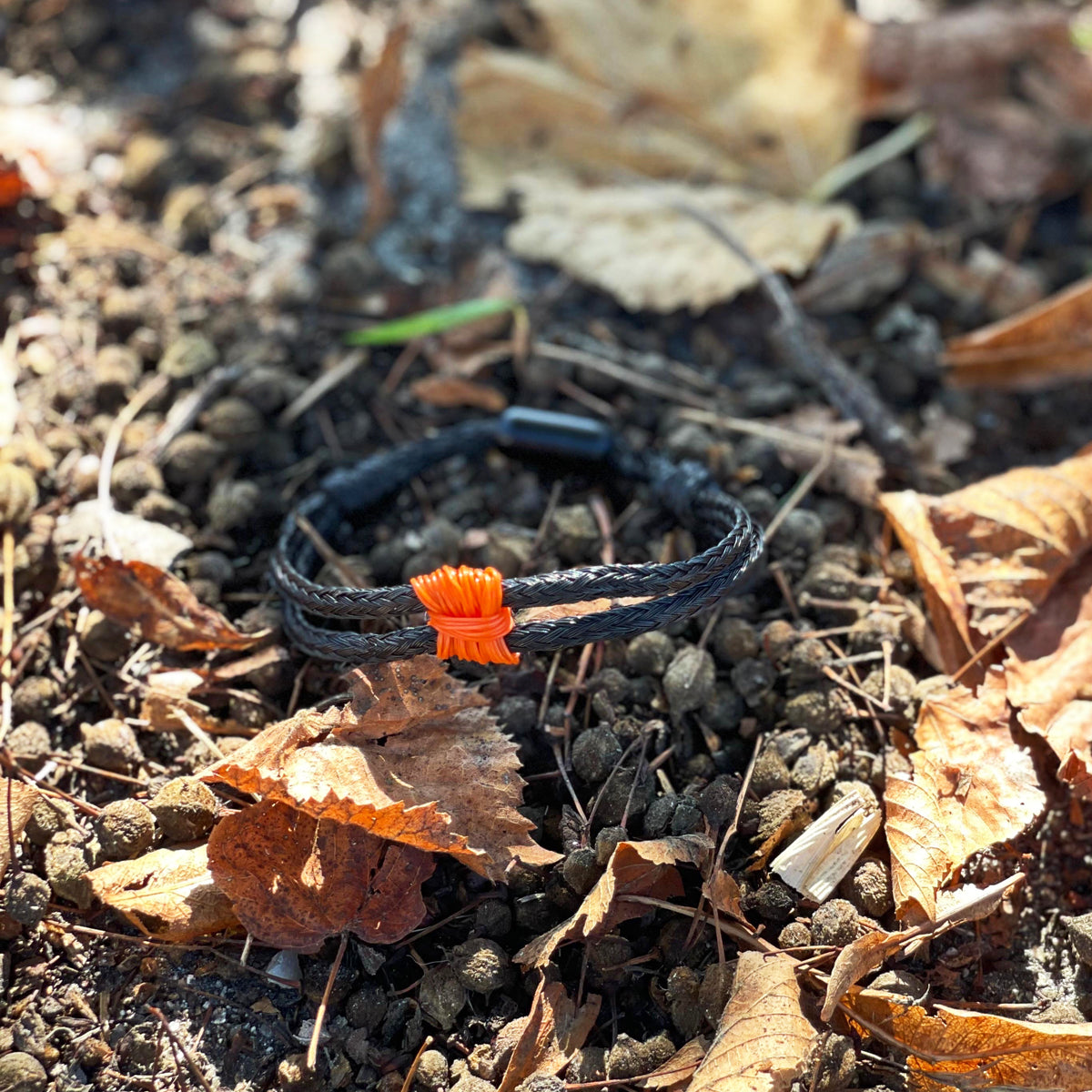Das BRACENET Upcycling Armband Pumpkin Bay II aus schwarzen alten Fischernetzen mit einem Knoten aus orangenen Dolly Ropes und einem schwarzen Magnetverschluss liegt auf der Erde. 