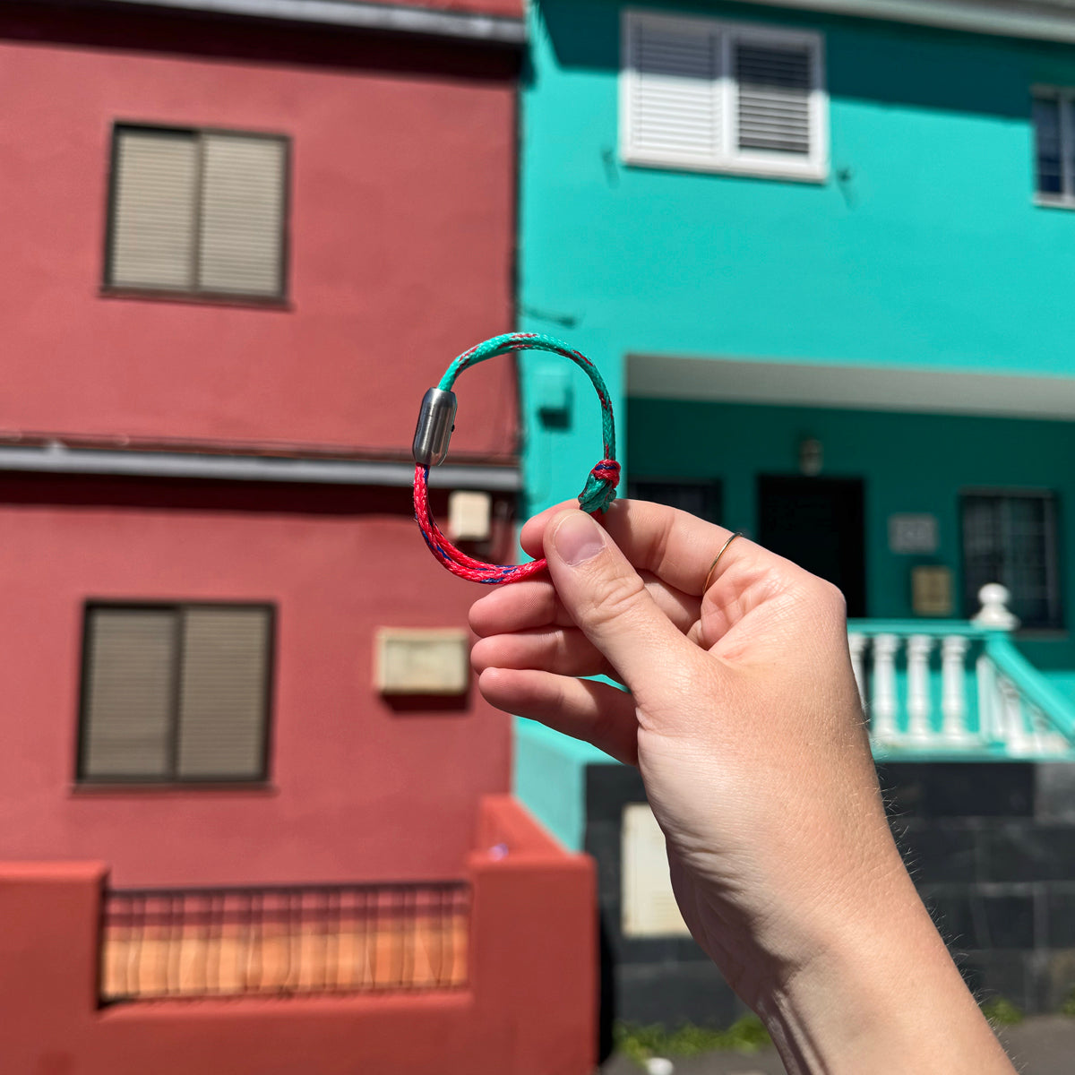 Person hält das April Bracenet Gulf of Mannar aus Fischernetz in Rot und Türkis mit Knoten und mit Bajonettverschluss in Silber vor zwei Häuser in den gleichen Farben.