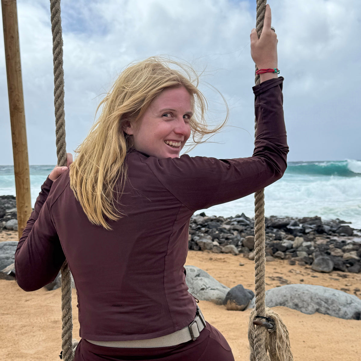 Person sitzt auf einer Schaukel am Meer und trägt das April Bracenet Gulf of Mannar aus Fischernetz in Rot und Türkis mit Knoten und mit Bajonettverschluss in Silber am Handgelenk. 