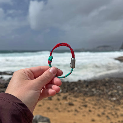 Person hält das April Bracenet Gulf of Mannar aus Fischernetz in Rot und Türkis mit Knoten und mit Bajonettverschluss in Silber vor das Meer. 