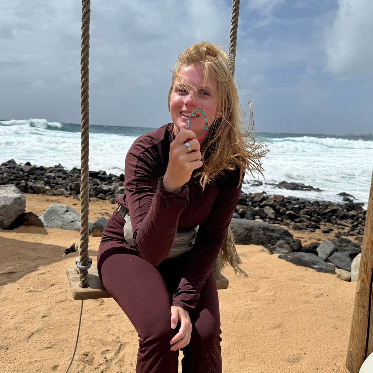 Person sitzt auf einer Schaukel am Meer und hält das April Bracenet Gulf of Mannar aus Fischernetz in Rot und Türkis mit Knoten und mit Bajonettverschluss in Silber vor das Gesicht. 