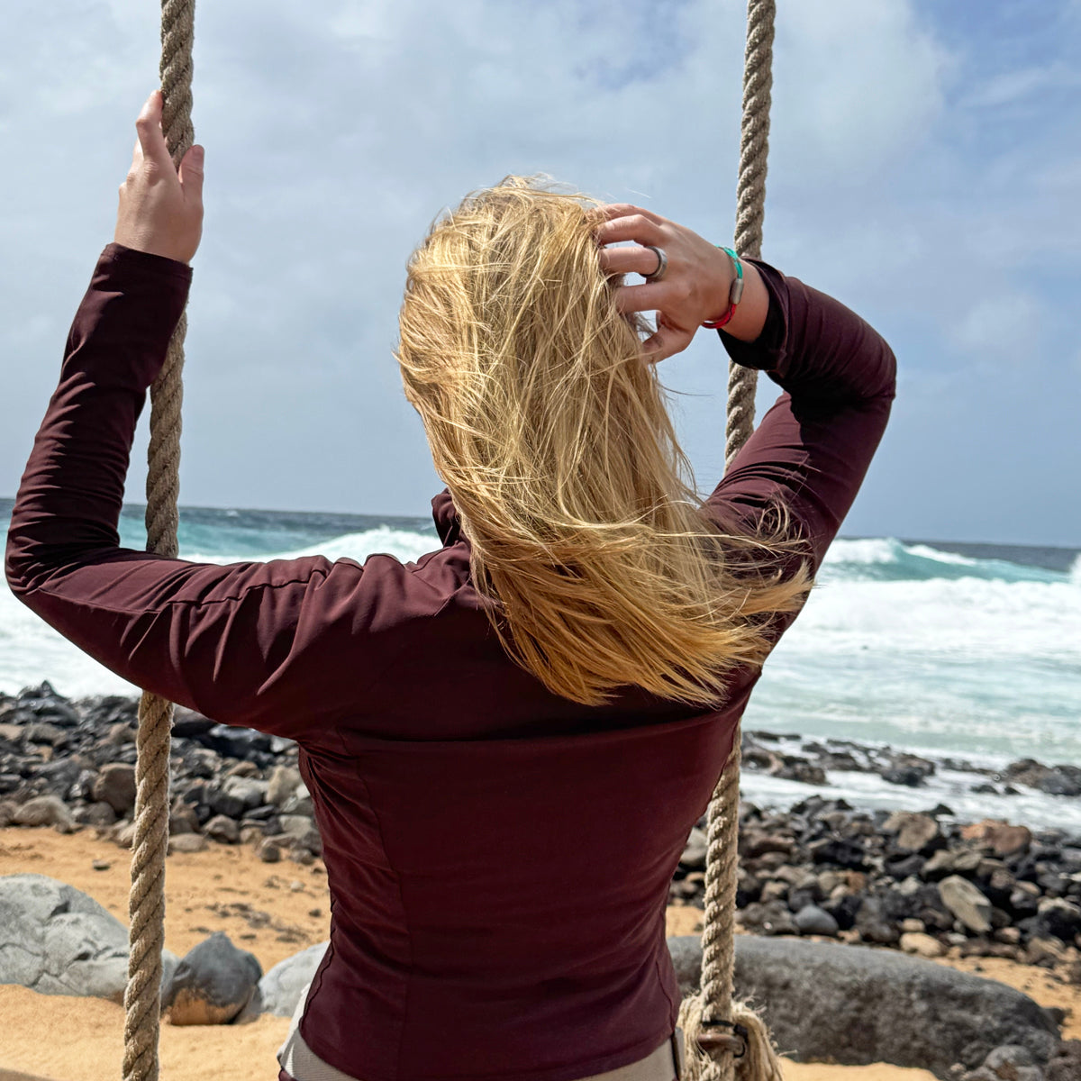 Person sitzt auf einer Schaukel am Meer, fasst sich durch die Haare und trägt das April Bracenet Gulf of Mannar aus Fischernetz in Rot und Türkis mit Knoten und mit Bajonettverschluss in Silber am Handgelenk. 