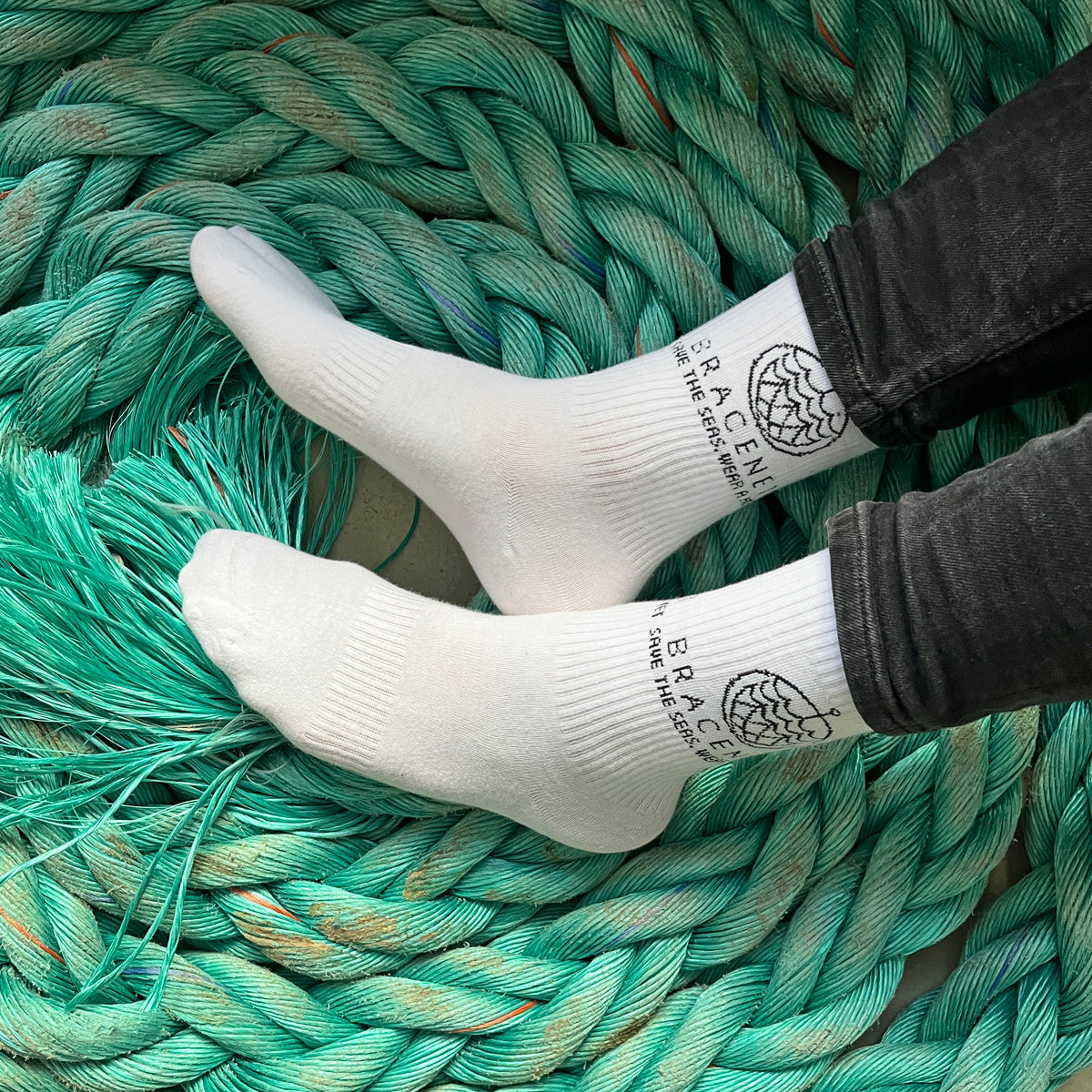 Person trägt BRACENET Baumwoll Socken in weiß mit schwarzem BRACENET Logo und sitzt auf einem alten Netz. 