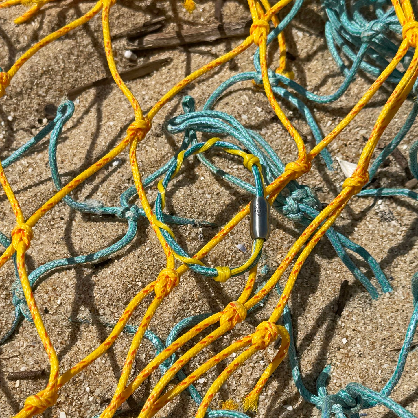 Mai Bracenet Mayotte's Lagoon aus Fischernetz in Hellblau und Gelb mit Knoten und mit Bajonettverschluss in Silber liegt am Sand auf Fischernetzen.