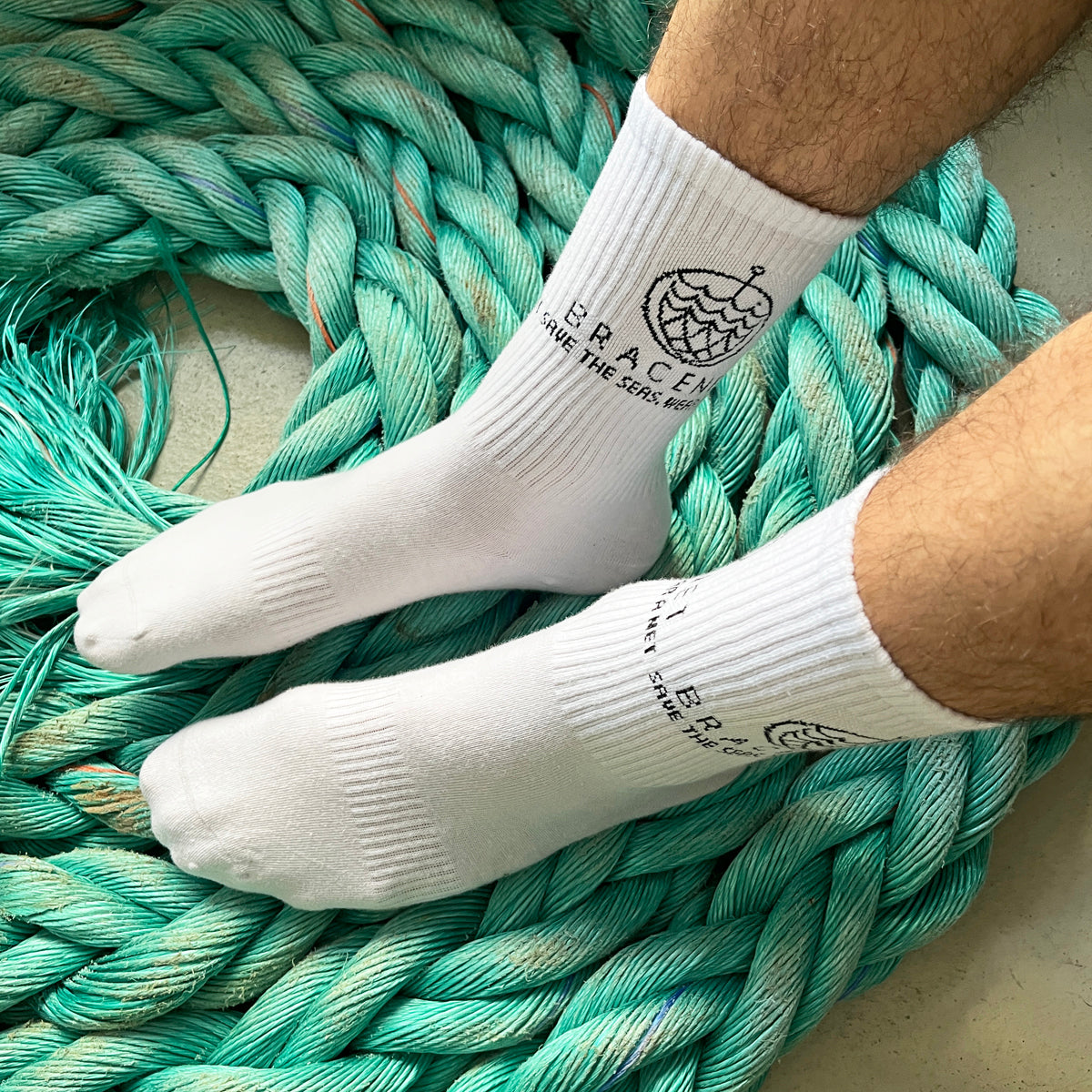 Person trägt BRACENET Baumwoll Socken in weiß mit schwarzem BRACENET Logo und sitzt auf einem alten Netz. 