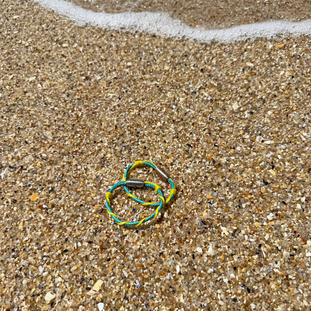 Mai Bracenet Mayotte's Lagoon aus Fischernetz in Hellblau und Gelb mit Knoten und mit Bajonettverschluss in Silber liegen im Sand am Meer. 