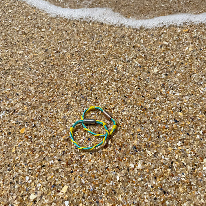 Mai Bracenet Mayotte's Lagoon aus Fischernetz in Hellblau und Gelb mit Knoten und mit Bajonettverschluss in Silber liegen im Sand am Meer. 