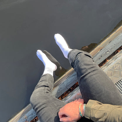 Person trägt BRACENET Baumwoll Socken in weiß mit schwarzem BRACENET Logo und sitzt auf einem Steg- Die Füße baumeln über dem Wasser. 
