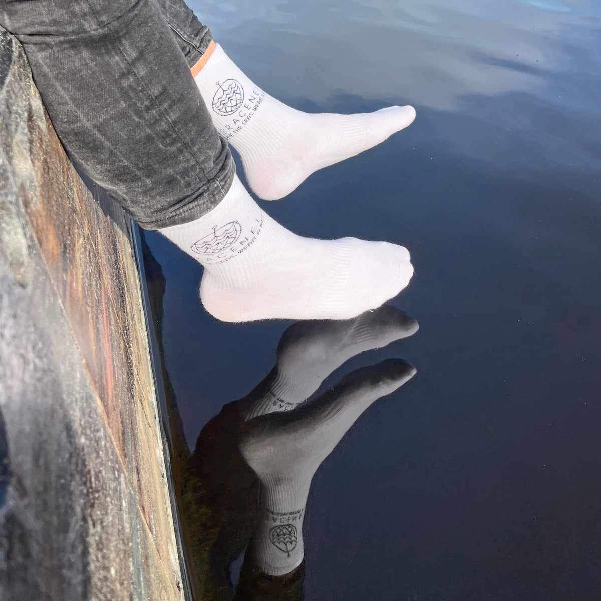 Person trägt BRACENET Baumwoll Socken in weiß mit schwarzem BRACENET Logo und sitzt auf einem Steg. Die Füße baumeln über dem Wasser. 