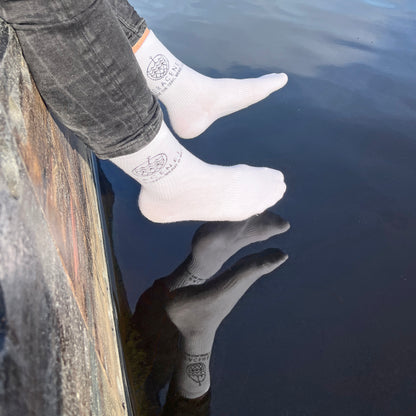 Person trägt BRACENET Baumwoll Socken in weiß mit schwarzem BRACENET Logo und sitzt auf einem Steg. Die Füße baumeln über dem Wasser. 