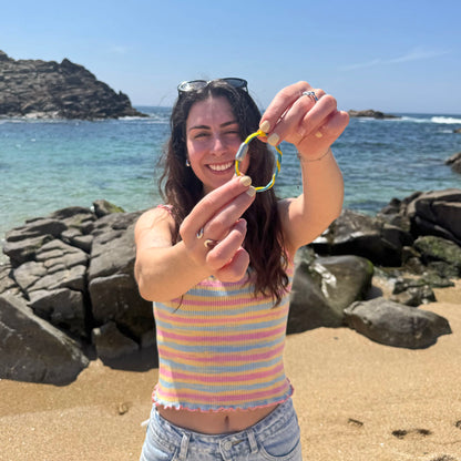 Person ist am Strand und hält das Mai Bracenet Mayotte's Lagoon aus Fischernetz in Hellblau und Gelb mit Knoten und mit Bajonettverschluss in Silber vors Gesicht. 