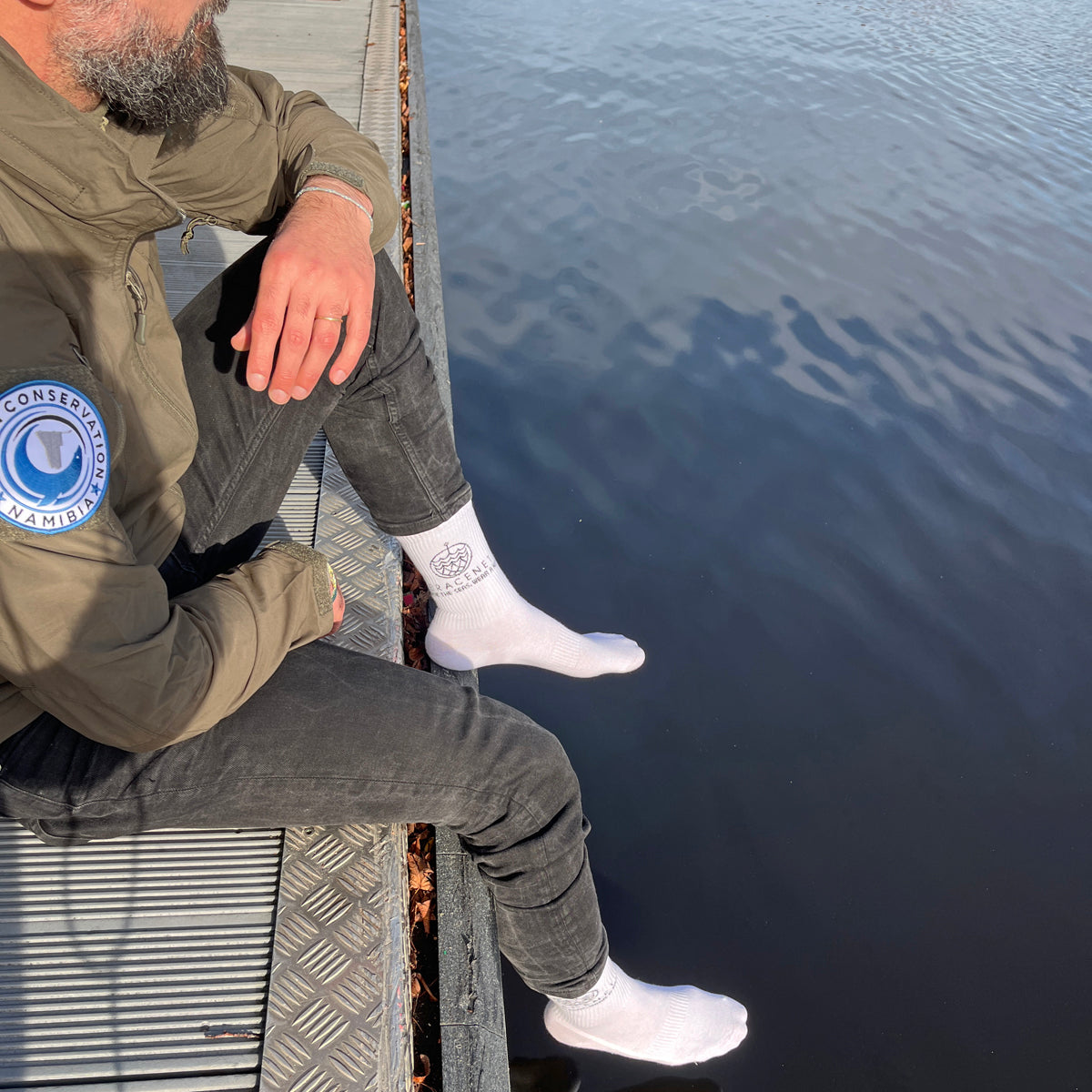 Person sitzt auf einem Steg am Wasser und trägt BRACENET Baumwoll Socken in weiß mit schwarzem BRACENET Logo.