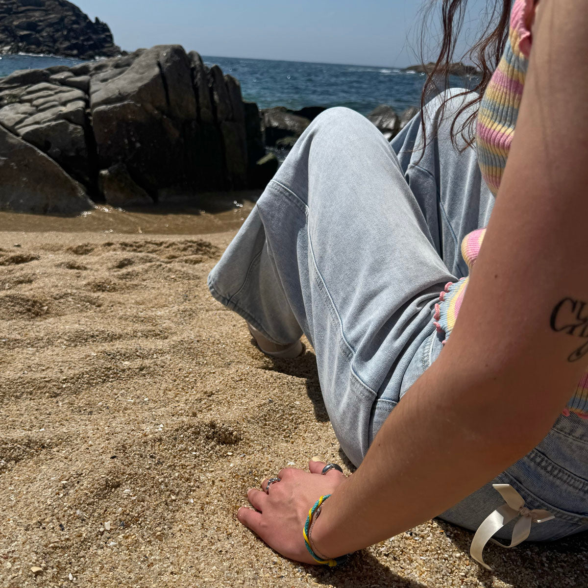 Person sitzt im Sand und hat das Mai Bracenet Mayotte's Lagoon aus Fischernetz in Hellblau und Gelb mit Knoten am Handgelenk. 