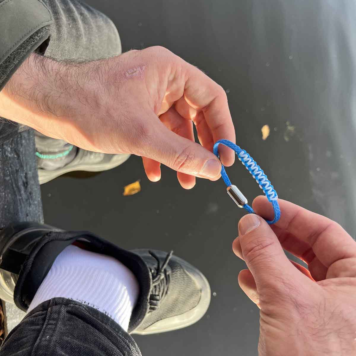 Person hält das Dezember BRACENET Upcycling Armband Arctic Ocean II aus blauen Fischernetzen mit Makramee Knoten in Hellblau und Magnetverschluss aus Edelstahl in Silber in den Händen über einer Wasseroberfläche. 