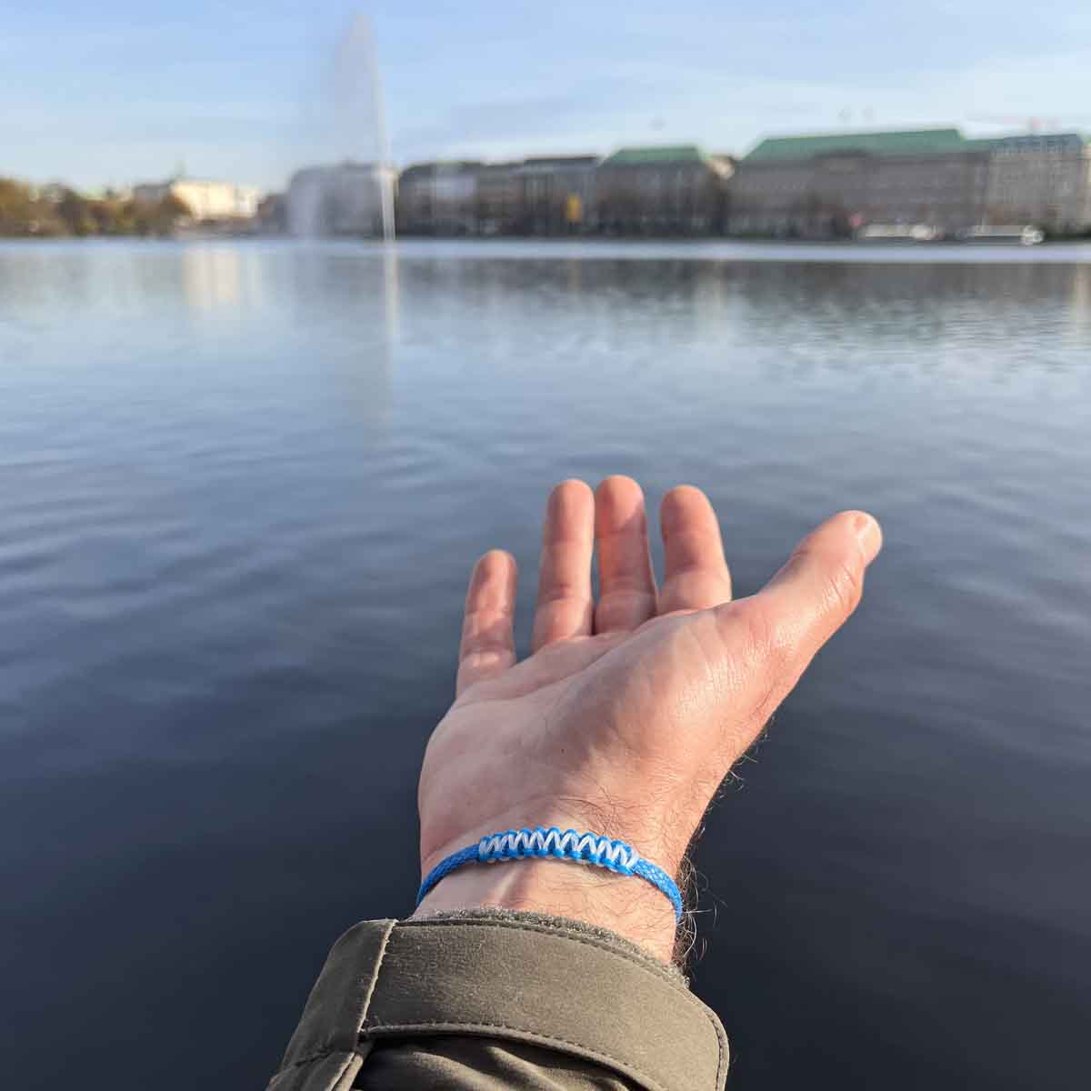 Person trägt das Dezember BRACENET Upcycling Armband Arctic Ocean II aus blauen Fischernetzen mit Makramee Knoten in Hellblau und Magnetverschluss aus Edelstahl in Silber am Handgelenk und  hält den Arm über das Wasser. 