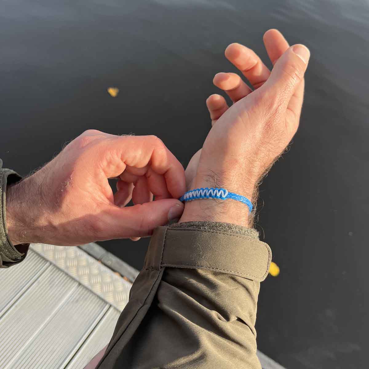 Person trägt Dezember BRACENET Upcycling Armband Arctic Ocean II aus blauen Fischernetzen mit Makramee Knoten in Hellblau und Magnetverschluss aus Edelstahl in Silber am Handgelenk und hält den Arm über eine Wasseroberfläche. 