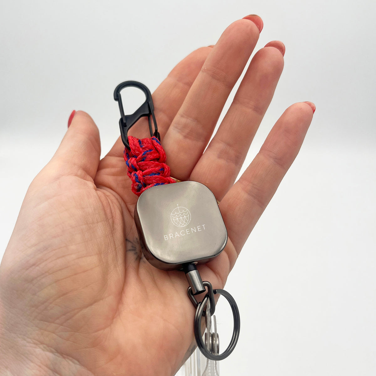Person hält den BRACENET Karten- und Schlüsselhalter mit ausziehbarer Zugrolle, BRACENET Logo, Schnürung aus Red Sea Fischernetz und schwarzem Karabiner in der Hand.