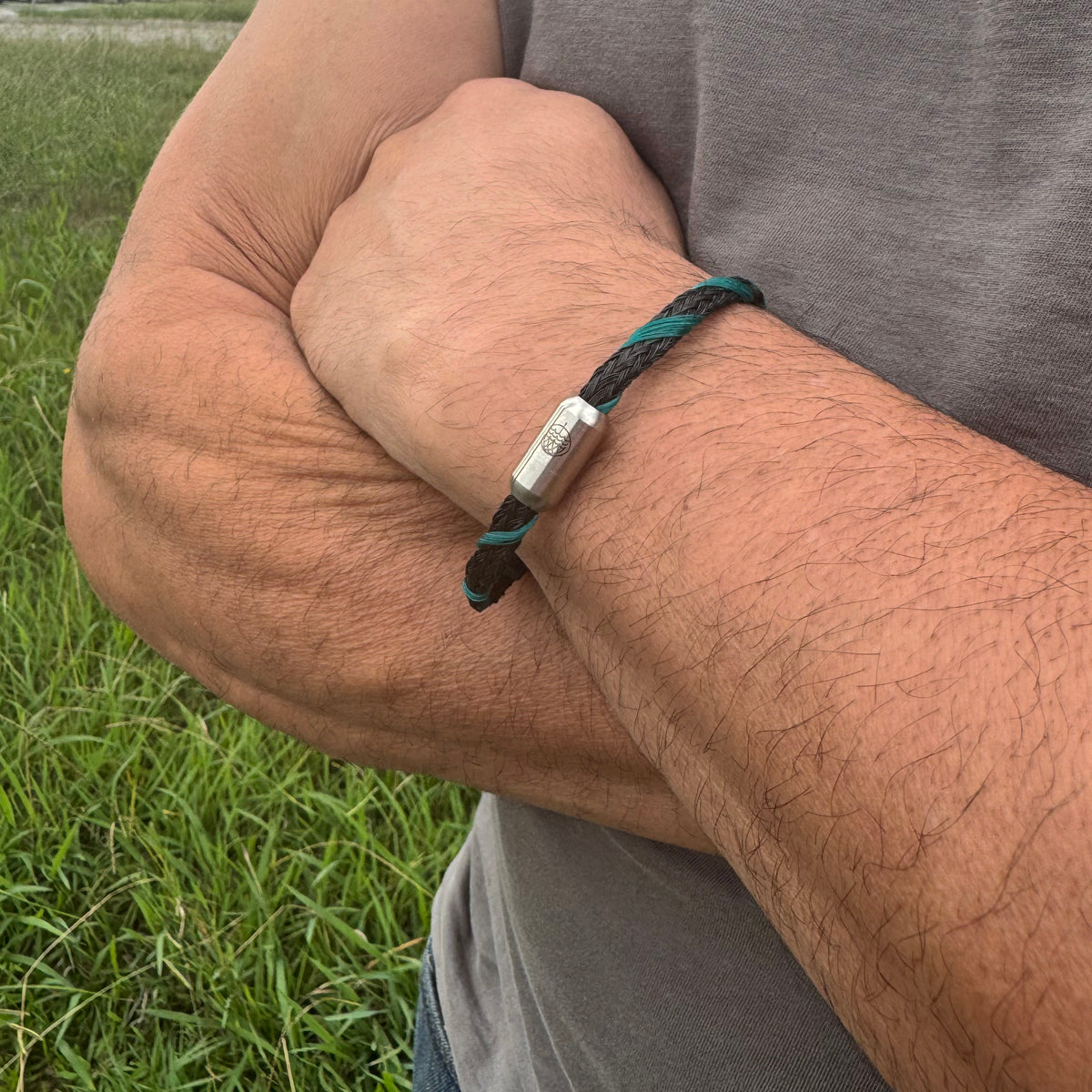Person trägt das November BRACENET Upcycling Armband Timor Sea aus schwarzen und petrolfarbenen Fischernetzen mit silber Verschluss am Handgelenk und hat die Arme verschränkt.