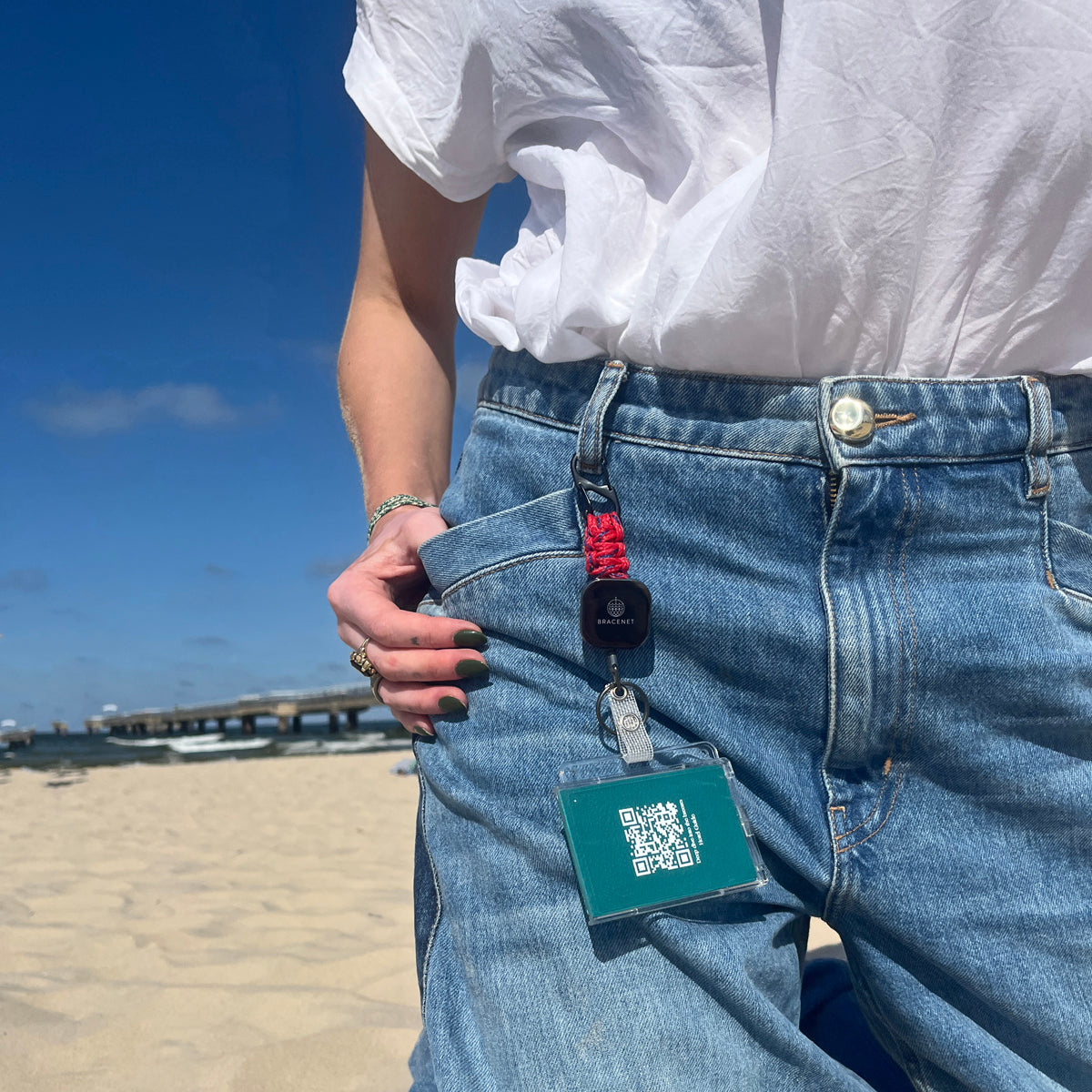 Person trägt den BRACENET Karten- und Schlüsselhalter mit ausziehbarer Zugrolle, BRACENET Logo, Schnürung aus Red Sea Fischernetz und schwarzem Karabiner an der Gürtelschlaufe einer Jeans und steht am Strand. 