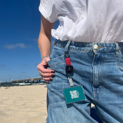 Person trägt den BRACENET Karten- und Schlüsselhalter mit ausziehbarer Zugrolle, BRACENET Logo, Schnürung aus Red Sea Fischernetz und schwarzem Karabiner an der Gürtelschlaufe einer Jeans und steht am Strand. 