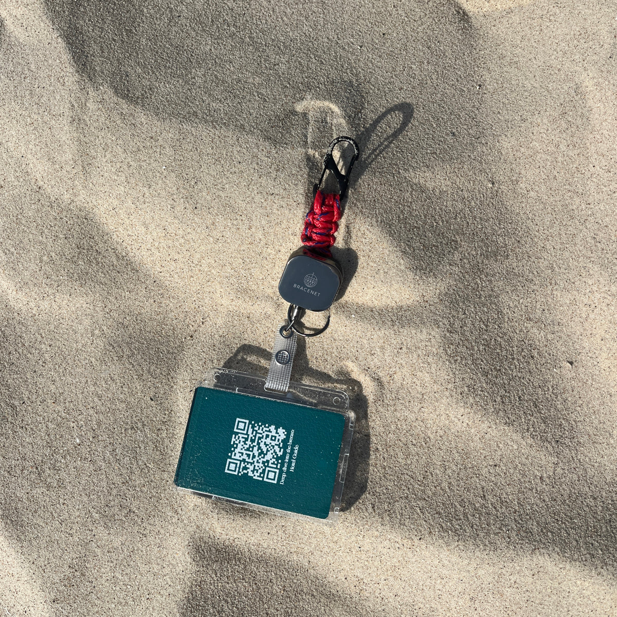 Der BRACENET Karten- und Schlüsselhalter mit ausziehbarer Zugrolle, BRACENET Logo, Schnürung aus Red Sea Fischernetz und schwarzem Karabiner liegt im Sand.