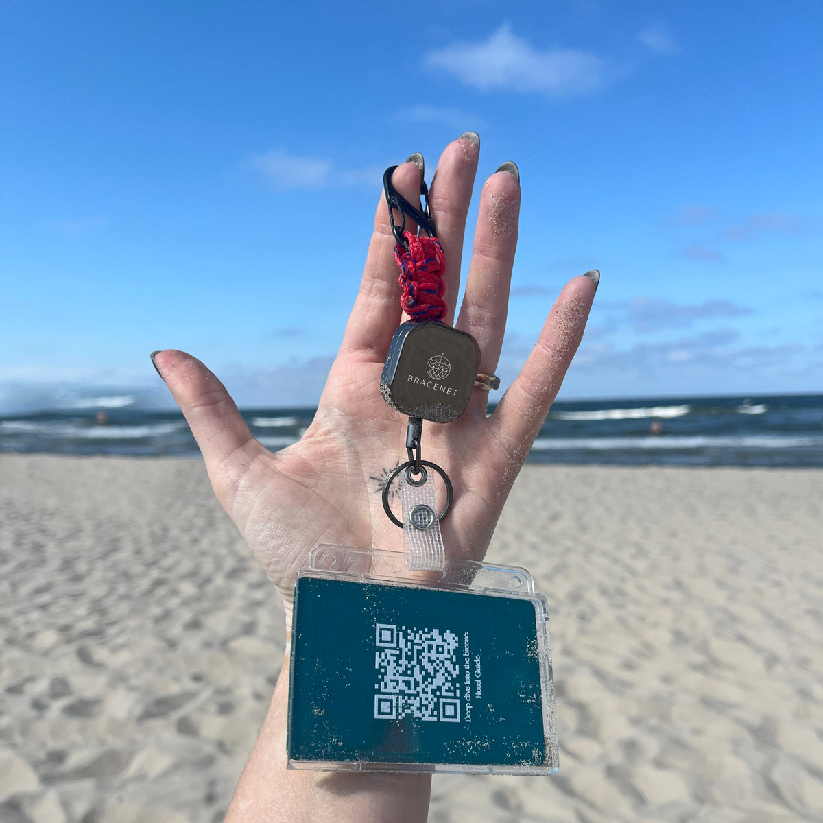 Person hält den BRACENET Karten- und Schlüsselhalter mit ausziehbarer Zugrolle, BRACENET Logo, Schnürung aus Red Sea Fischernetz und schwarzem Karabiner in der Hand vor sich. Im Hintergrund sind man das Meer. 